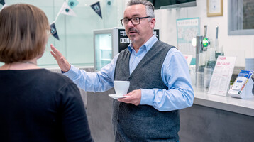 Theatre guest wearing a shirt and waistcoat standing in the Box Office Cafe holding a cup of coffee and talking to a member of staff