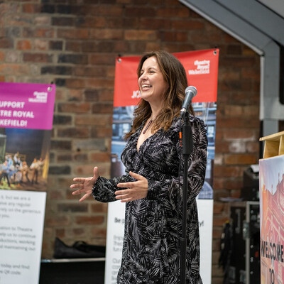 One woman wearing a patterned dress standing by a microphone and presenting to the side. Theatre Royal Wakefield banners in the background.