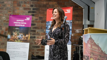 One woman wearing a patterned dress standing by a microphone and presenting to the side. Theatre Royal Wakefield banners in the background.