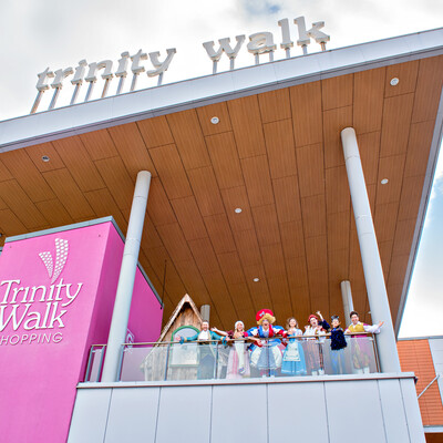 Image from below of shopping centre building with large signs reading 'Trinity Walk'. Cast all dressed in pantomime costumes standing on edge looking down at camera.