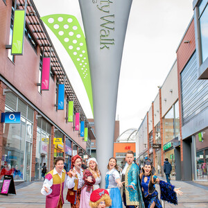 Cast all dressed in pantomime costumes posing around a 'Trinity Walk' sign, with shops in the background.