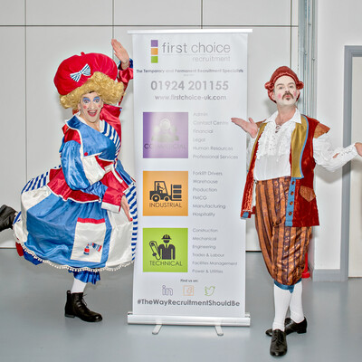 Pantomime dame and male character both in costume, posing and pointing to 'First Choice Recruitment' banner.