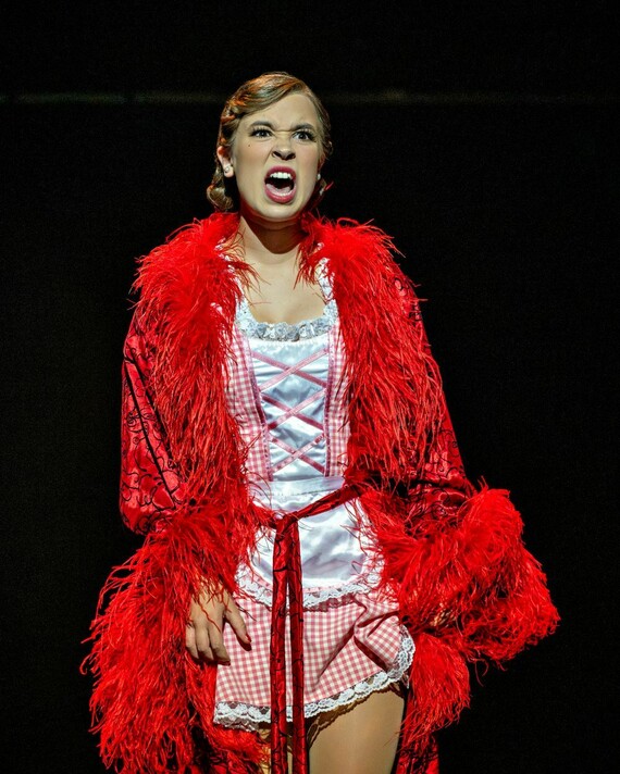 Miss Adelaide is wearing a bright red feathered coat and red and white gingham dress. She has an angry expression.