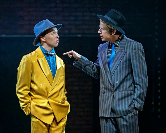 Nathan Detroit wearing a yellow suit, blue shirt, and blue hat. Sky Masterson is pointing at Nathan in a black and white pinstripe suit, striped shirt, blue tie, and black hat. Both men have one hand in their trouser pockets.