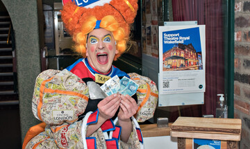 Photo of Chris Hannon dressed as Winnie Whittington holding up £5 notes next to the donation box by the auditorium.