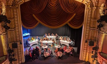 A view of the stage taken from the Upper Circle. The stage is filled with tables and chairs, with diners enjoying a three course meal.