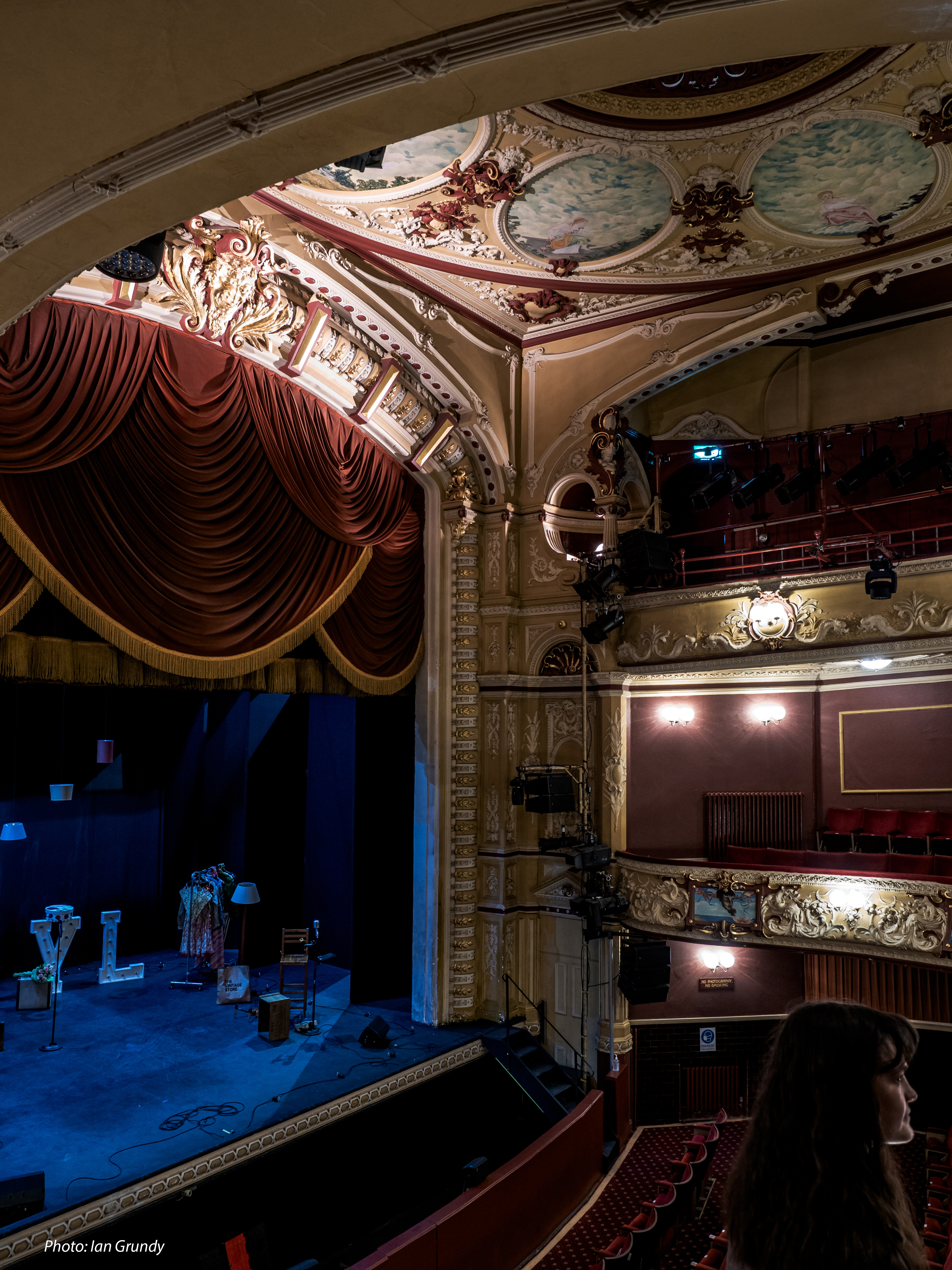 Photo of the stage in the Matcham Auditorium. Decorative painted ceiling and proscenium arch stage with red curtains and seating.